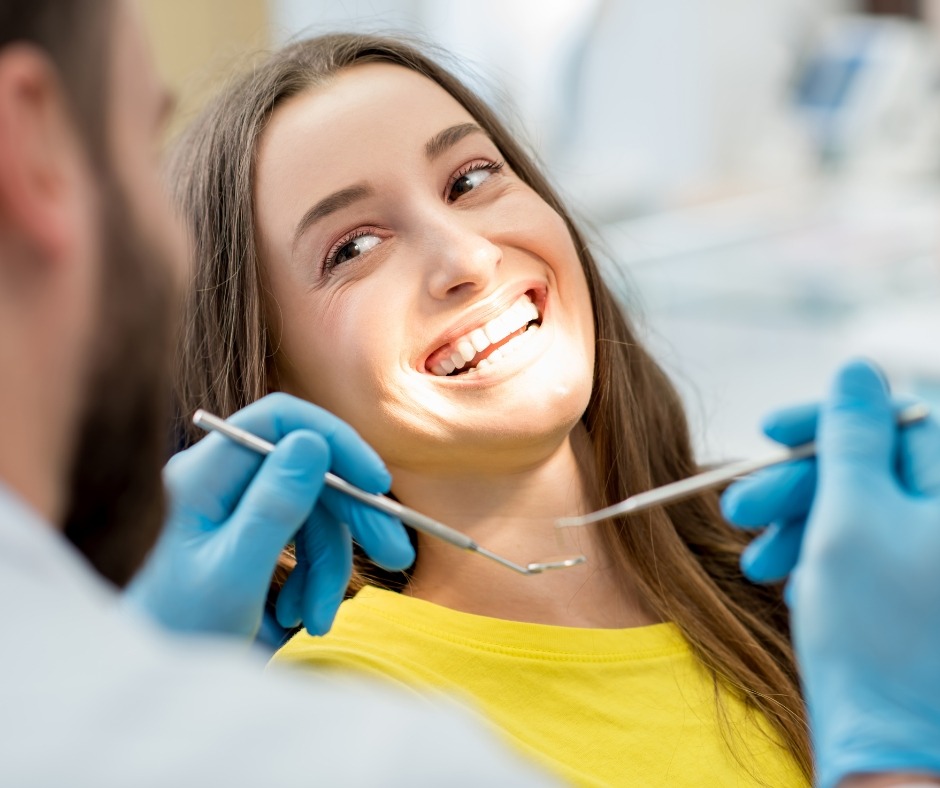 A woman smiling with a dentist holding some dental tools in the foreground.