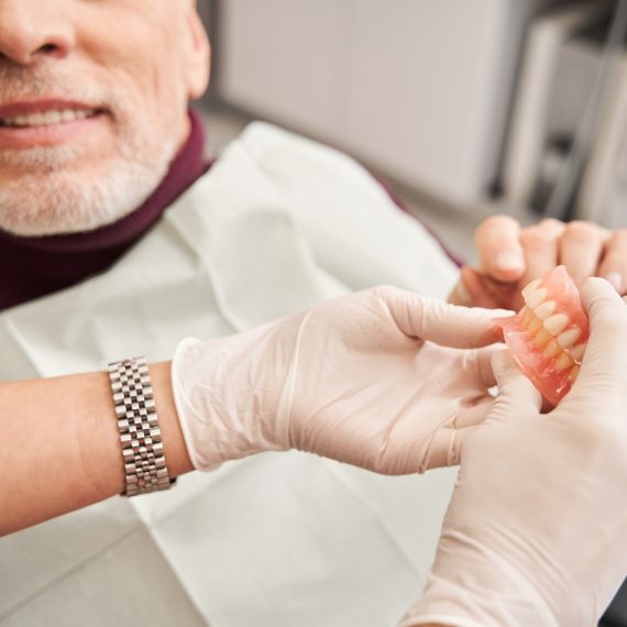 View of the dentist showing to senior grey haired patient teeth dentures while working at the dental clinic.