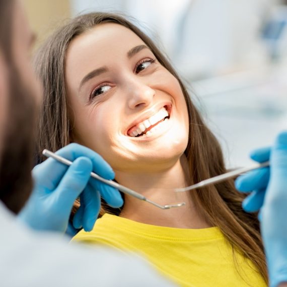 A woman smiling with a dentist holding some dental tools in the foreground.